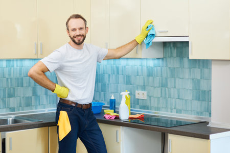 Housekeeping services. Cheerful professional male cleaner wearing gloves smiling at camera while cleaning kitchen cabinets indoorsの写真素材