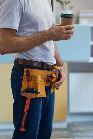 Coffee break. Cropped shot of professional repairman wearing a tool belt taking a break, drinking coffee while standing indoorsの写真素材