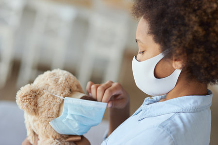 Mixed race teen girl child in protective face mask holding a teddy bear wearing a maskの写真素材