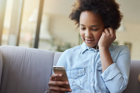 Portrait of pretty mixed race teen girl putting on earphones, sitting on the couch and using mobile phone while spending time at homeの写真素材