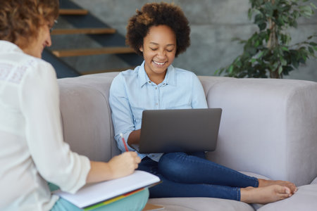 Cute teen girl smiling, using laptop for studying while having a lesson with her female teacher, sitting on the sofa at homeの写真素材