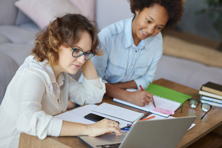 Portrait of focused female teacher in glasses helping teen schoolgirl with homework, sitting at the table at homeの写真素材