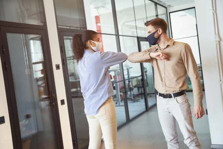Covid 19 infection prevention. Two young diverse colleagues wearing face protective masks bumping elbows, greeting each other while standing in the modern officeの写真素材