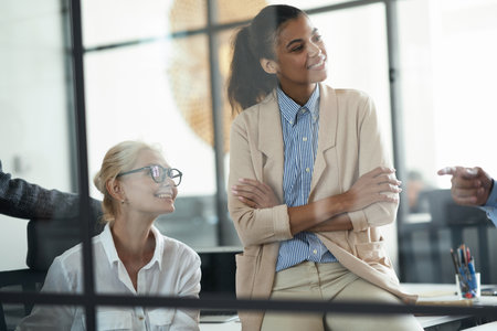 Two smiling diverse female coworkers looking aside while having a meeting with colleagues in the modern officeの写真素材