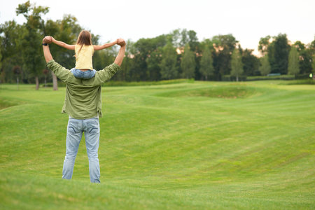 Rear view of little girl sitting on fathers shoulders and holding each other hands, standing in a green field on a warm day, spending time together outdoorsの写真素材