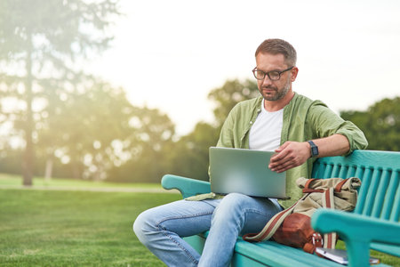 Young man wearing eyeglasses looking focused while working using laptop, sitting on the bench in the green parkの写真素材