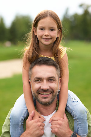 Portrait of happy dad holding his cute little daughter on shoulders and smiling at camera while having fun together in the green park on a warm dayの写真素材