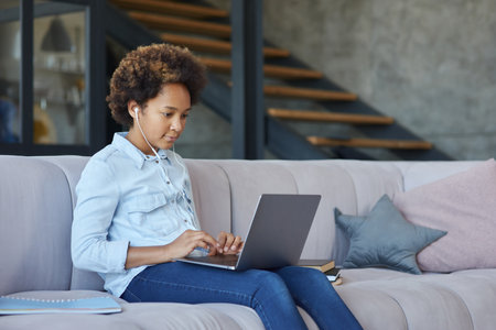 Cute teen schoolgirl wearing earphones looking focused while using laptop, chatting with friends or teacher during online lesson, sitting on the couch at homeの写真素材