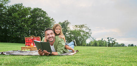Cheerful cute little girl spending time with her father, reading a book while having picnic in the parkの写真素材