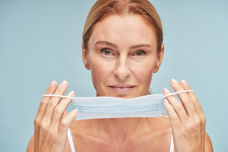 Portrait of adult beautiful lady holding medical mask in front of face isolated on blue backgroundの写真素材