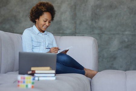 Cute mixed race teen schoolgirl smiling while doing homework, sitting with laptop and books on the couch at homeの写真素材