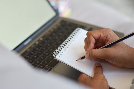 Close up shot of mixed race teen schoolgirl using pencil and making notes while doing homework, sitting with laptop at homeの写真素材