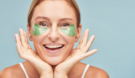 Beauty treatment. Portrait of happy caucasian mature woman with hydrogel patches under eyes smiling at camera isolated on blue backgroundの写真素材
