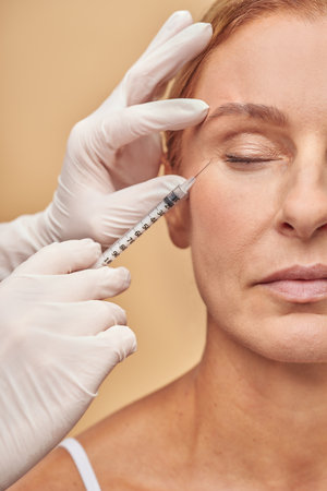 Vertical shot of beautician hands in white protective gloves with syringe making cosmetic injection for mature womanの写真素材