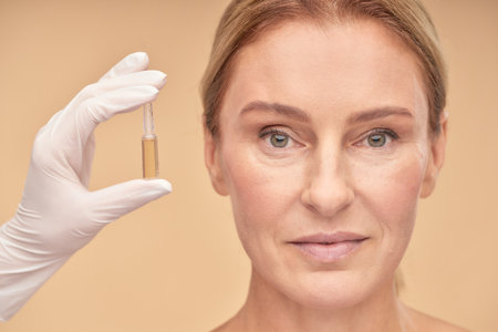 Cosmetology and skincare. Beautician hand in white glove holding ampoule with moisturizing face serum near mature woman faceの写真素材