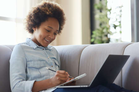 Explore and discover. Joyful mixed race teen schoolgirl using pencil for making notes while doing homework, sitting with laptop on the couch at homeの写真素材