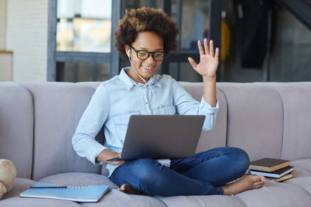 Happy teen schoolgirl wearing earphones looking cheerful while using laptop, greeting friends and teacher during online video lesson, sitting on the couch at homeの写真素材