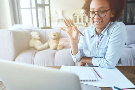 Joyful mixed race teen girl in glasses raised her hand while having online lesson via video chat app using laptop, studying in the living room at homeの写真素材