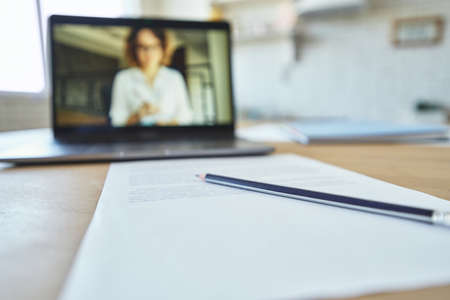 Close up of pencil and sheet of paper on the table. Laptop screen with excited female teacher communicating during video call in the backgroundの写真素材