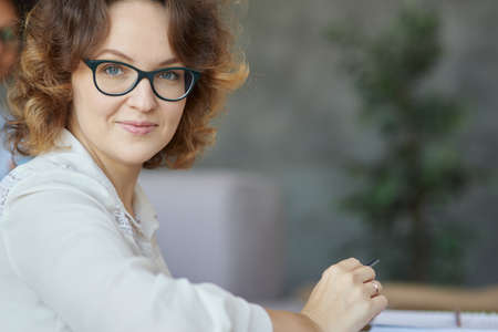 Kind teacher. Portrait of attractive young female teacher in glasses smiling at camera while having a lesson at homeの写真素材
