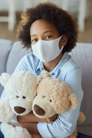 Portrait of cute mixed race teen girl in casual clothes wearing protective face mask, looking at camera, hugging her toys while sitting on a sofa at homeの写真素材