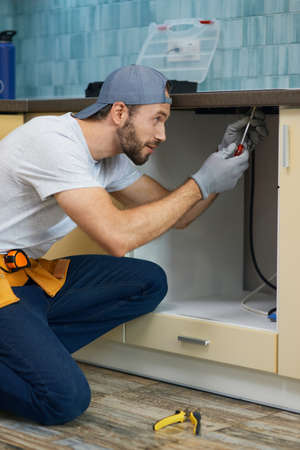 Fixing leaks. Focused young repairman, professional plumber wearing tool belt crouching on the floor while fixing sink pipe in the kitchen indoorsの写真素材