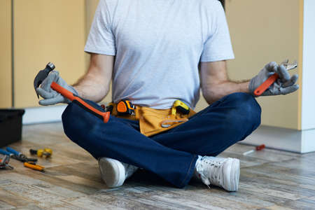 Cropped shot of young handyman holding a hammer and an adjustable wrench in his hands while meditating, sitting in Lotus pose on the floor indoorsの写真素材