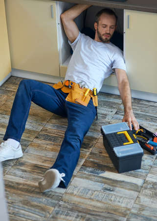 Plumbing expert. Professional repairman, plumber wearing tool belt examining and fixing sink pipe in the kitchen indoorsの写真素材