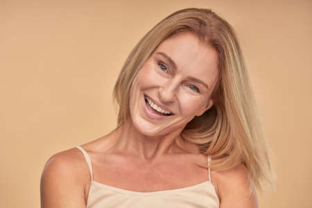 Flirty beauty. Portrait of beautiful and happy mature woman looking aside and laughing while posing in studio over beige backgroundの写真素材