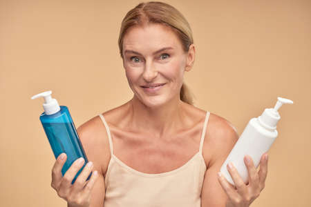 Portrait of a beautiful mature woman choosing between two cosmetic products while standing isolated over beige background in studioの写真素材