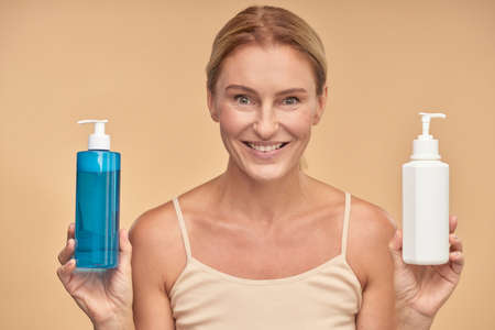 Portrait of a happy woman choosing between two body care products while standing isolated over beige background in studioの写真素材