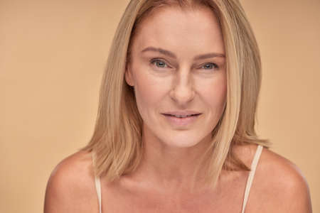 Close up portrait of beautiful caucasian mature woman looking at camera while posing in studio over beige backgroundの写真素材