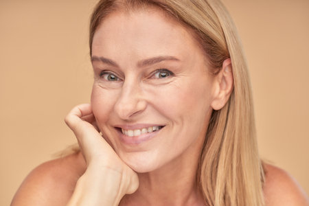 Anti-aging treatments. Close up portrait of attractive happy middle aged woman touching her face and smiling at camera while posing over beige background in studioの写真素材