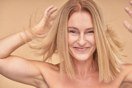 Feeling joyful. Portrait of happy attractive mature woman playing with her hair and smiling at camera while standing over beige backgroundの写真素材