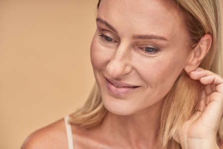 Portrait of charming middle aged caucasian woman with clean and soft skin touching her hair, looking aside and smiling while posing over beige background in studioの写真素材
