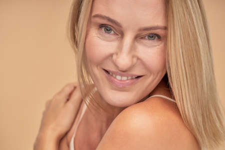 Portrait of beautiful adult woman touching her shoulders with her hands and looking at camera while posing in studio over beige backgroundの写真素材