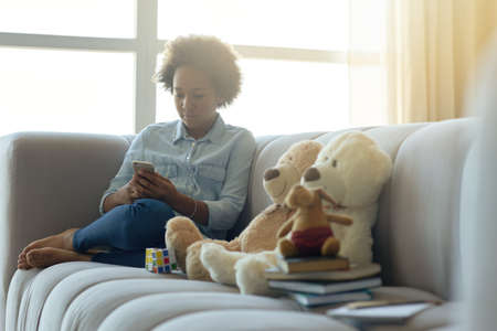 Modern childhood. Cheerful mixed race teen girl sitting on the couch and using mobile phone while spending time at homeの写真素材