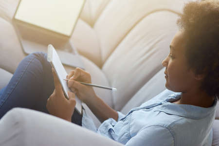 Close up shot of mixed race teen schoolgirl using pencil for making notes while doing homework, sitting on the couch at homeの写真素材