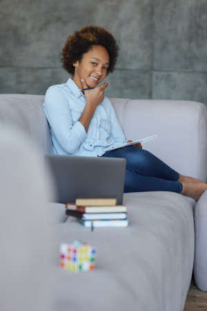 Cute mixed race teen schoolgirl smiling at camera while doing homework, sitting with laptop and books on the couch at homeの写真素材