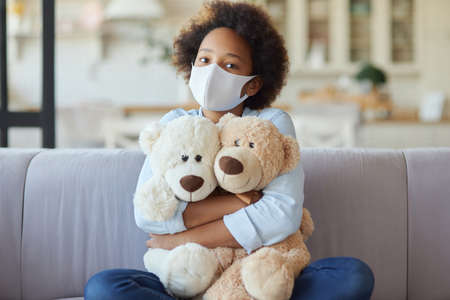 Portrait of cute mixed race teen girl in casual clothes wearing protective face mask, looking at camera, hugging her toys while sitting on a sofa at homeの写真素材