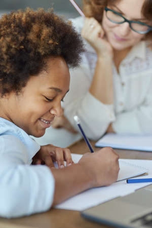 Close up shot of enthusiastic mixed race teen schoolgirl writing with pencil during a lesson with her female teacher at homeの写真素材