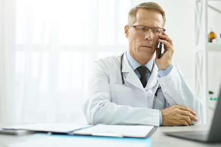 Serious doctor talking on mobile phone and using laptop at workの写真素材
