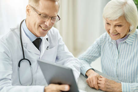 Cheerful doctor using tablet computer while talking with old ladyの写真素材