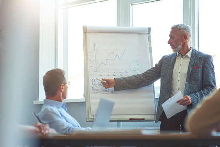 Confident mature businessman pointing at flip chart and explaining something to colleagues while...