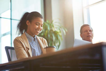 Female office worker. Young happy mixed race woman using laptop and smiling while sitting with male colleague at desk in the modern officeの写真素材