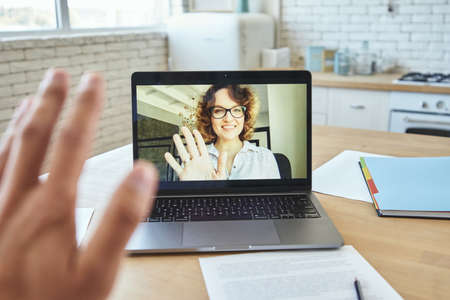 Close up of laptop screen with attractive female teacher smiling and waving at student during video call, having online lesson during quarantine lockdownの写真素材