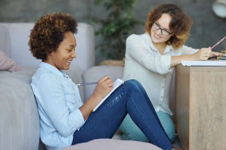 Joyful mixed race teen schoolgirl smiling while making notes during a lesson with her female teacher, sitting on the couch at homeの写真素材