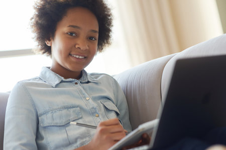 Cute mixed race teen schoolgirl smiling at camera while doing homework, sitting with laptop on the couch at homeの写真素材