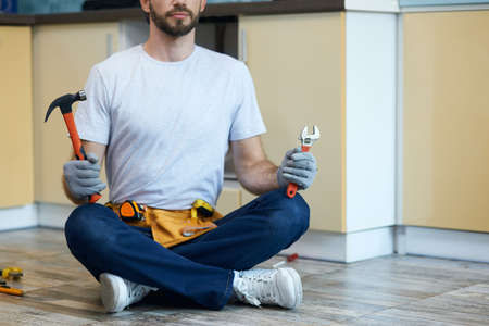 Serving you. Cropped shot of young handyman holding a hammer and an adjustable wrench in his hands while sitting on the floor indoorsの写真素材