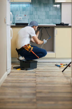 Pipes need to be maintained. Young repairman, professional plumber wearing tool belt crouching on the floor while fixing sink pipe in the kitchen indoorsの写真素材